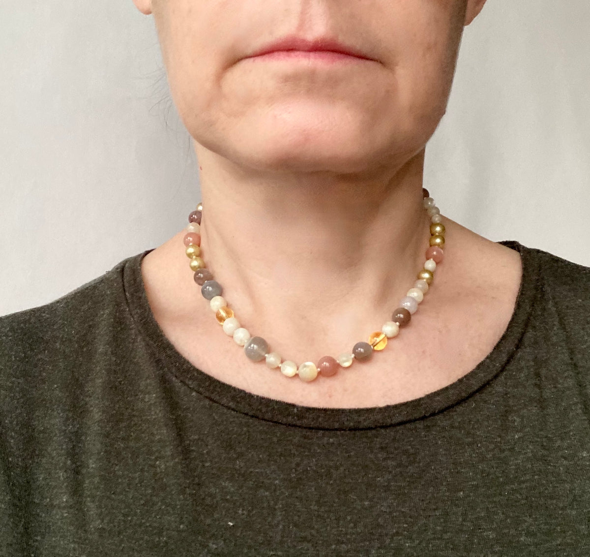 A close-up of a person wearing a multi-colored beaded necklace with gray, cream, orange, and gold-toned beads, paired with a dark olive green shirt.