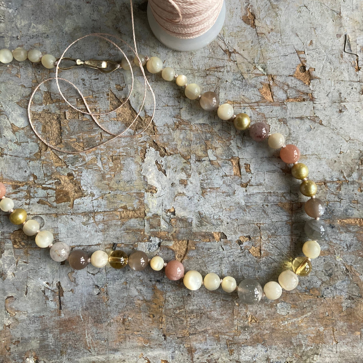 A beaded jewelry-making strand with round beads in cream, gray, peach, and metallic gold, laid out on a distressed gray wooden surface next to a spool of light pink thread.