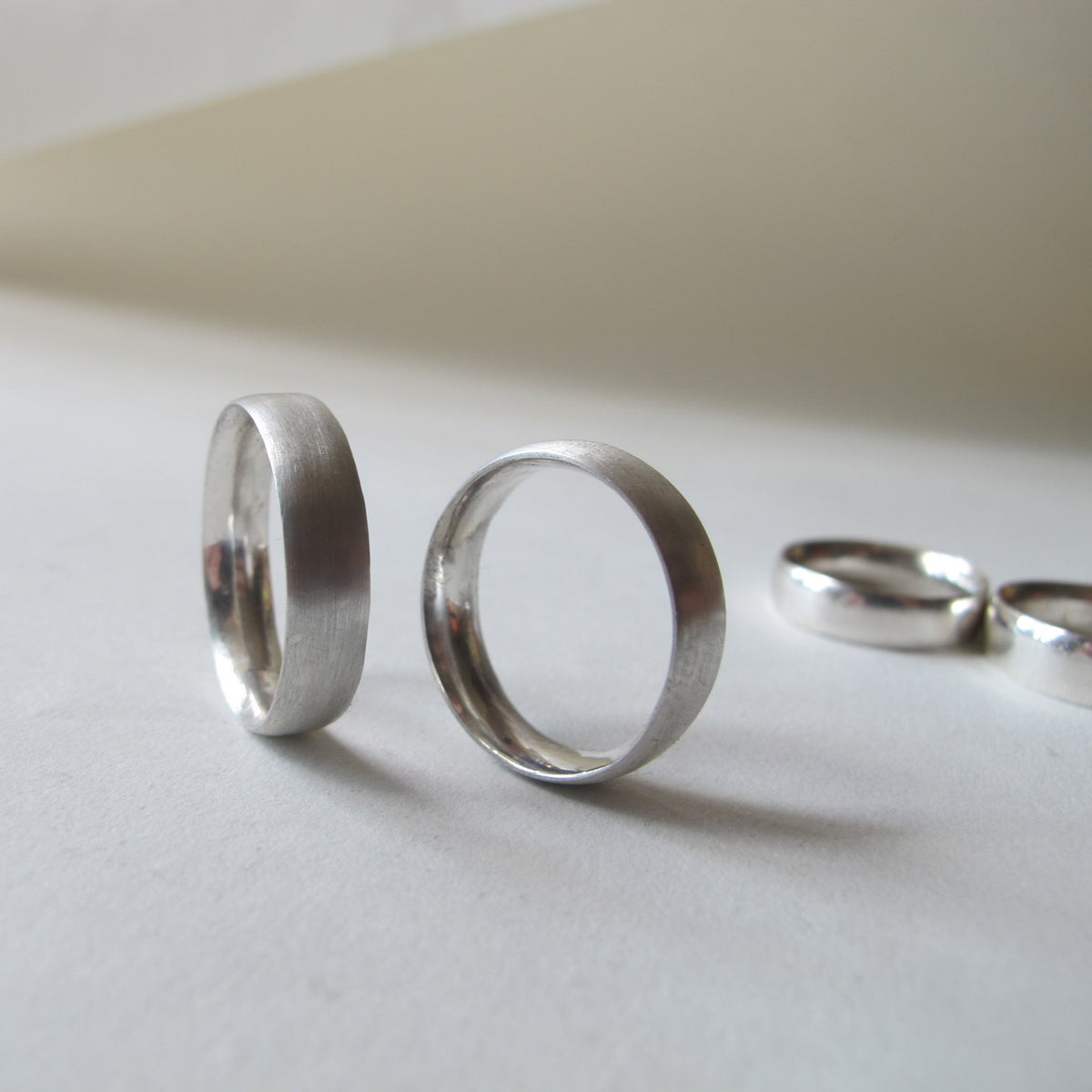 Two silver metal bands, one with a brushed matte finish and one with a polished satin finish, displayed on a light gray surface with other rings blurred in the background.