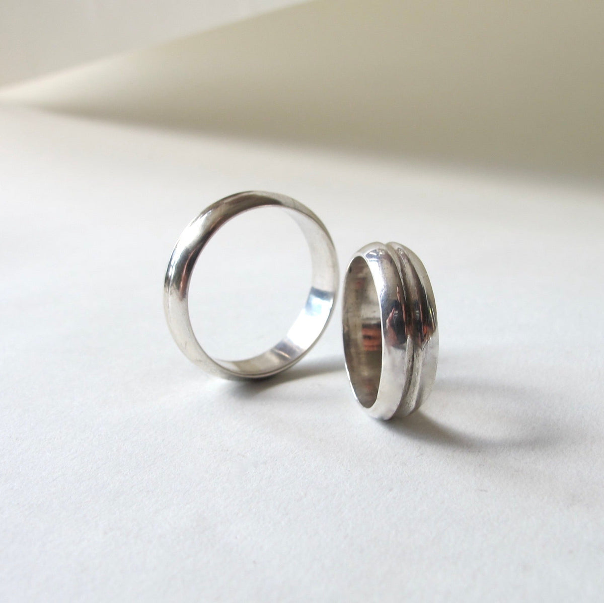 Two polished silver rings, one with a smooth matte finish and one with a textured ridged band, displayed on a light off-white surface.