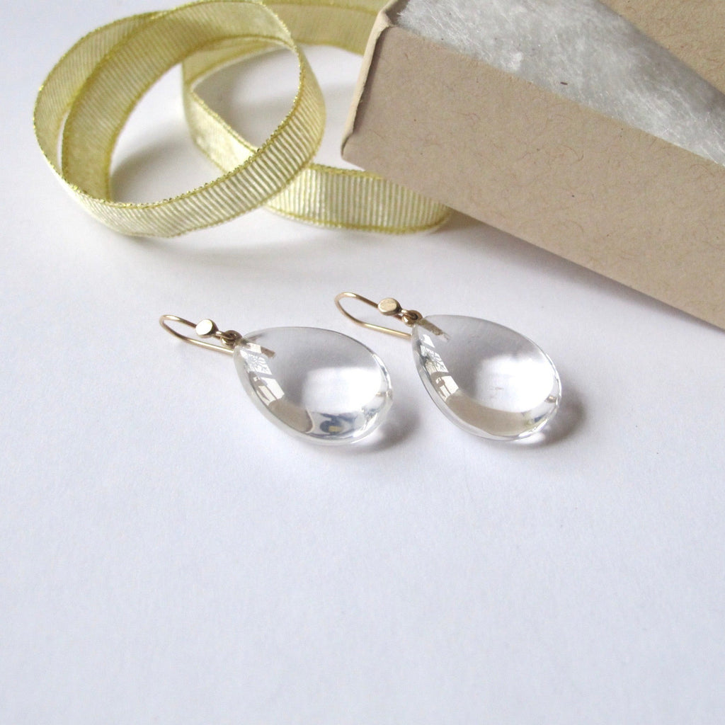 Clear teardrop-shaped glass or acrylic drop earrings with gold fishhook clasps, shown beside a gold ribbon and a brown kraft gift box on a white background.