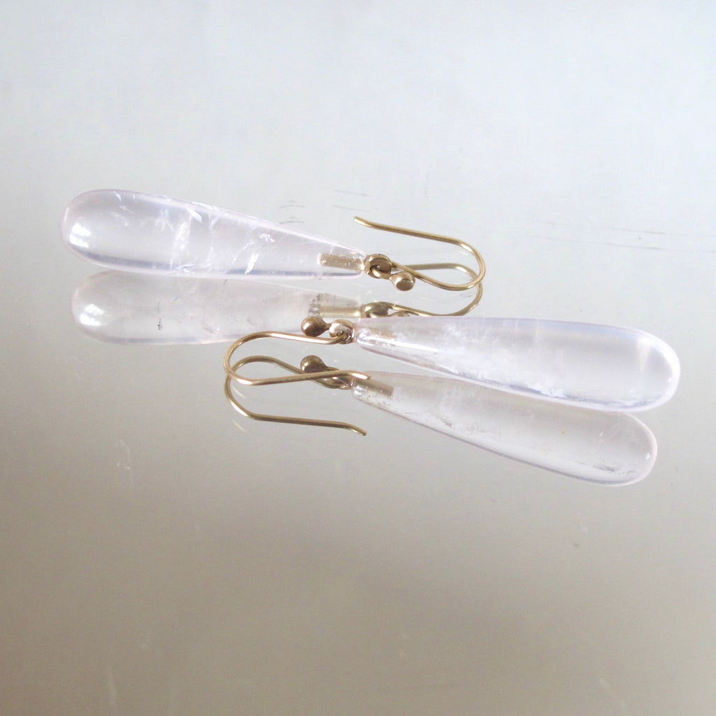 A pair of clear resin teardrop dangle earrings with gold fishhook ear wires, displayed on a light beige reflective surface.