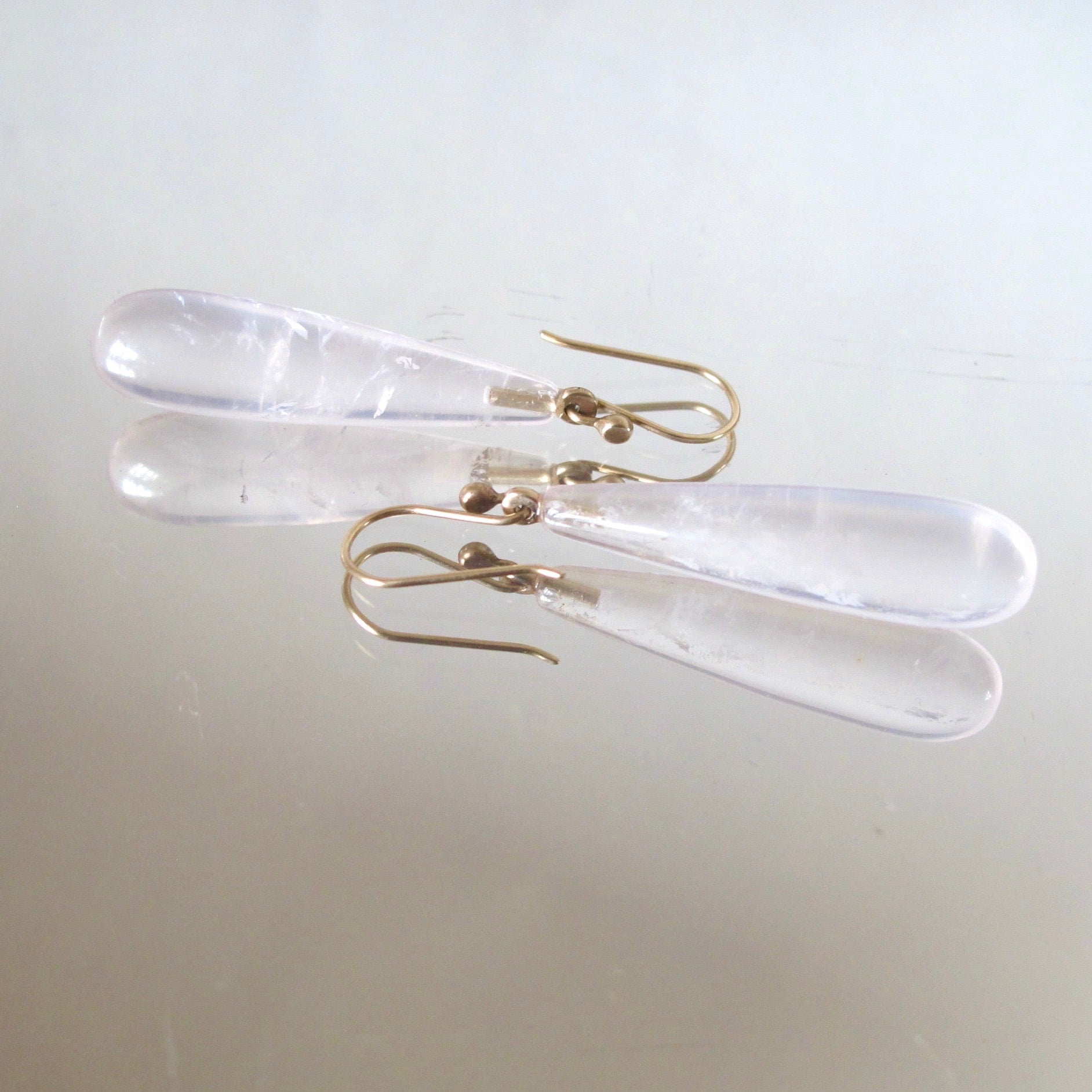 A pair of clear resin teardrop dangle earrings with gold fishhook ear wires, displayed on a light beige reflective surface.