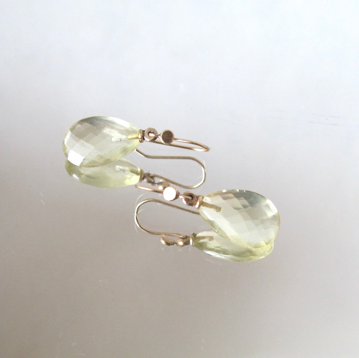 Faceted light green teardrop earrings with gold fishhook wires, displayed on a reflective light gray surface.