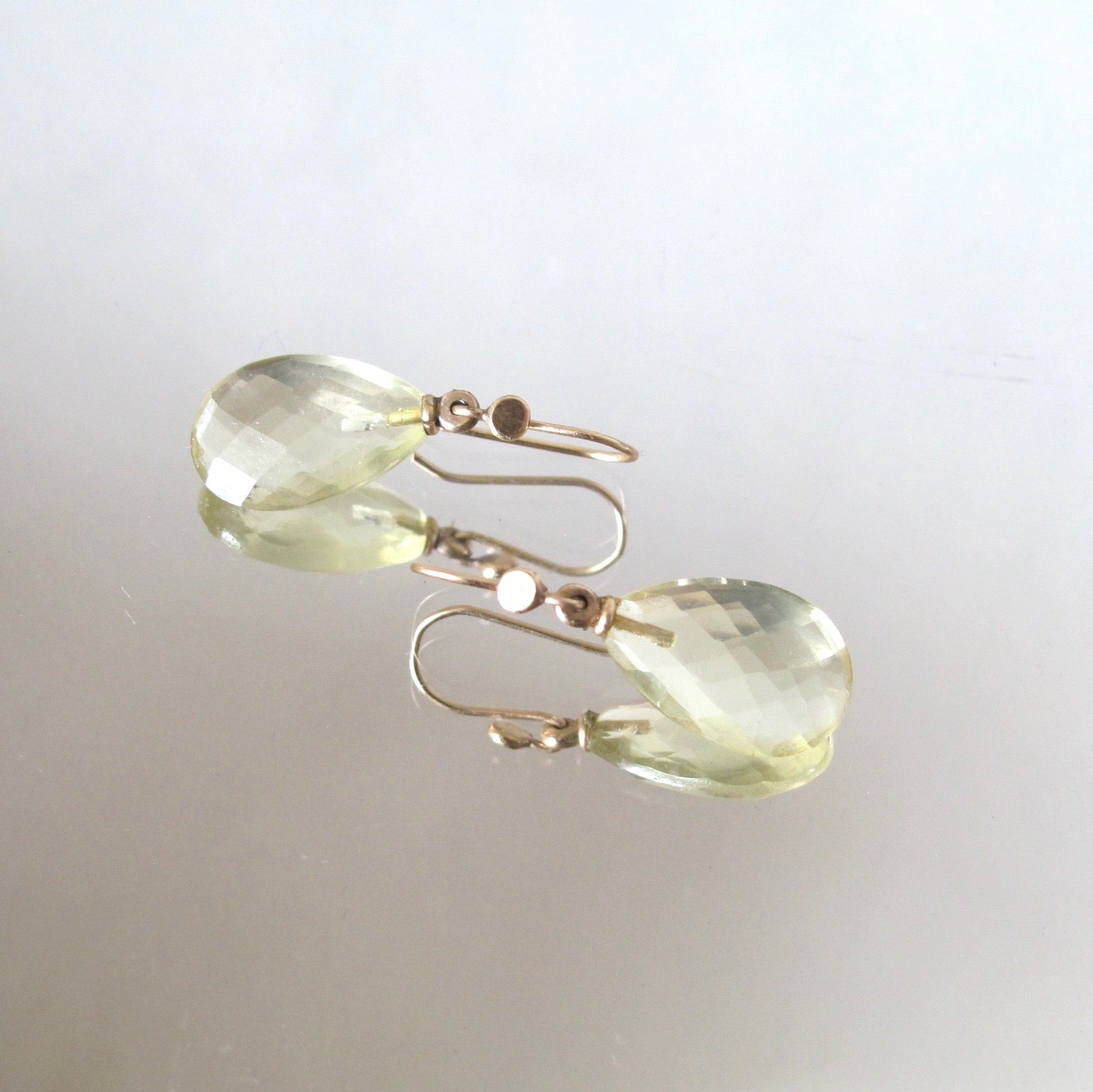 Faceted light green teardrop earrings with gold fishhook wires, displayed on a reflective light gray surface.
