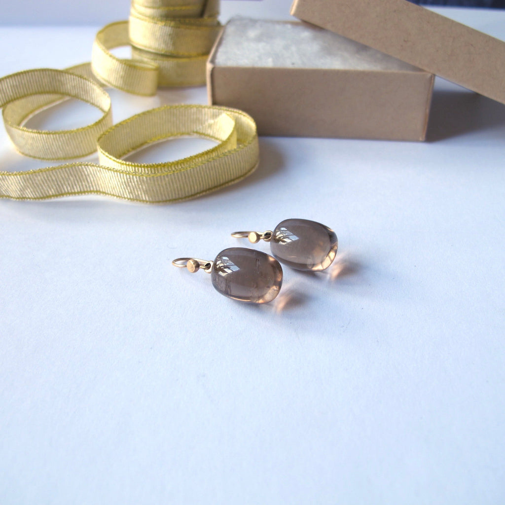 A pair of gold drop earrings with oval smoky brown glass beads, displayed on a white surface next to a roll of yellow ribbon and a brown kraft gift box.
