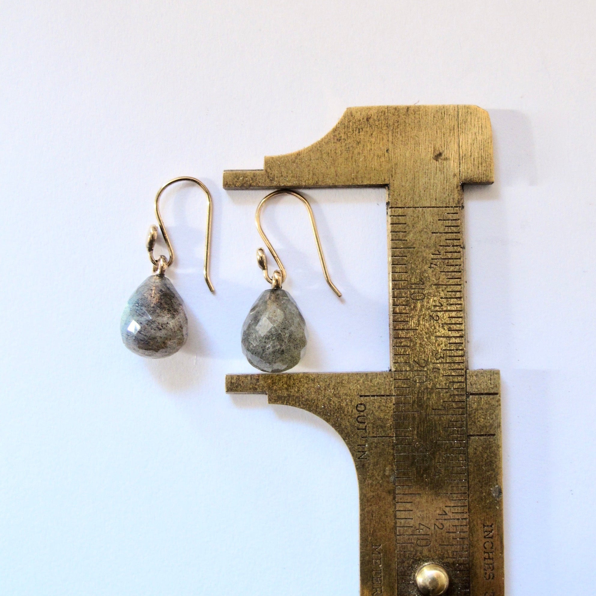 A pair of teardrop-shaped dangle earrings with metallic gold fishhook wires and gray-green marbled stone drops, shown next to a brass caliper against a plain white background.