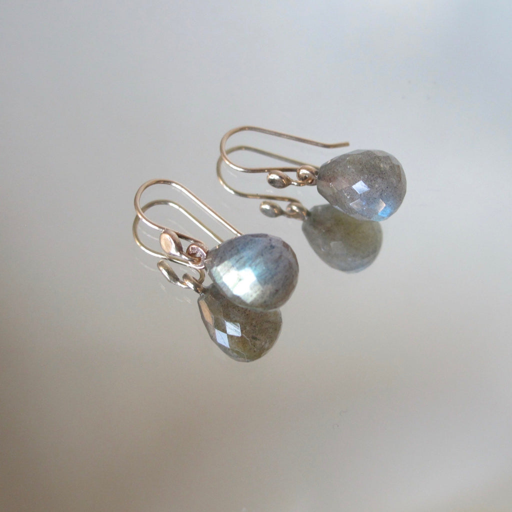 A pair of teardrop-shaped labradorite drop earrings with rose gold hooks, displayed on a reflective surface showing their mirror image.