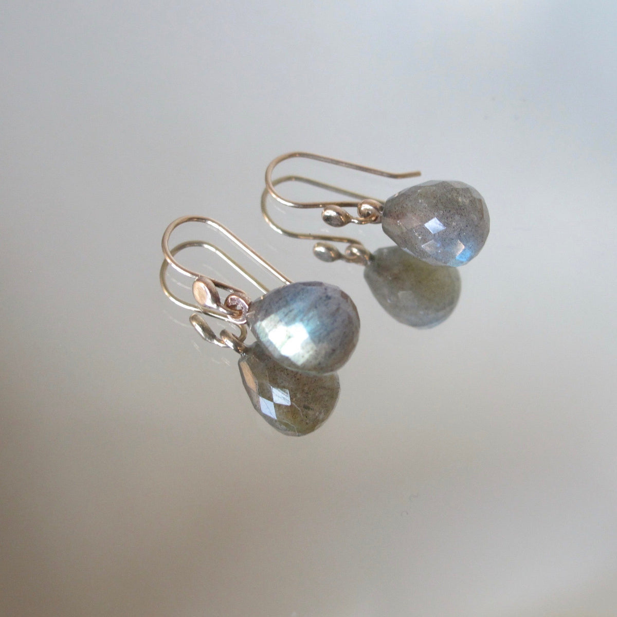 A pair of teardrop-shaped labradorite drop earrings with rose gold hooks, displayed on a reflective surface showing their mirror image.