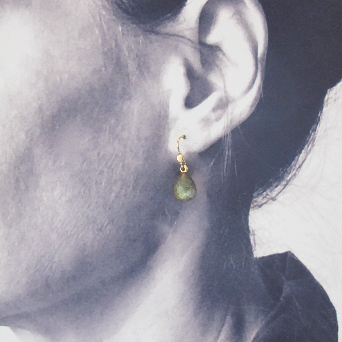 A close-up side profile of a person wearing a small gold hoop earring with a teardrop-shaped green stone dangle.