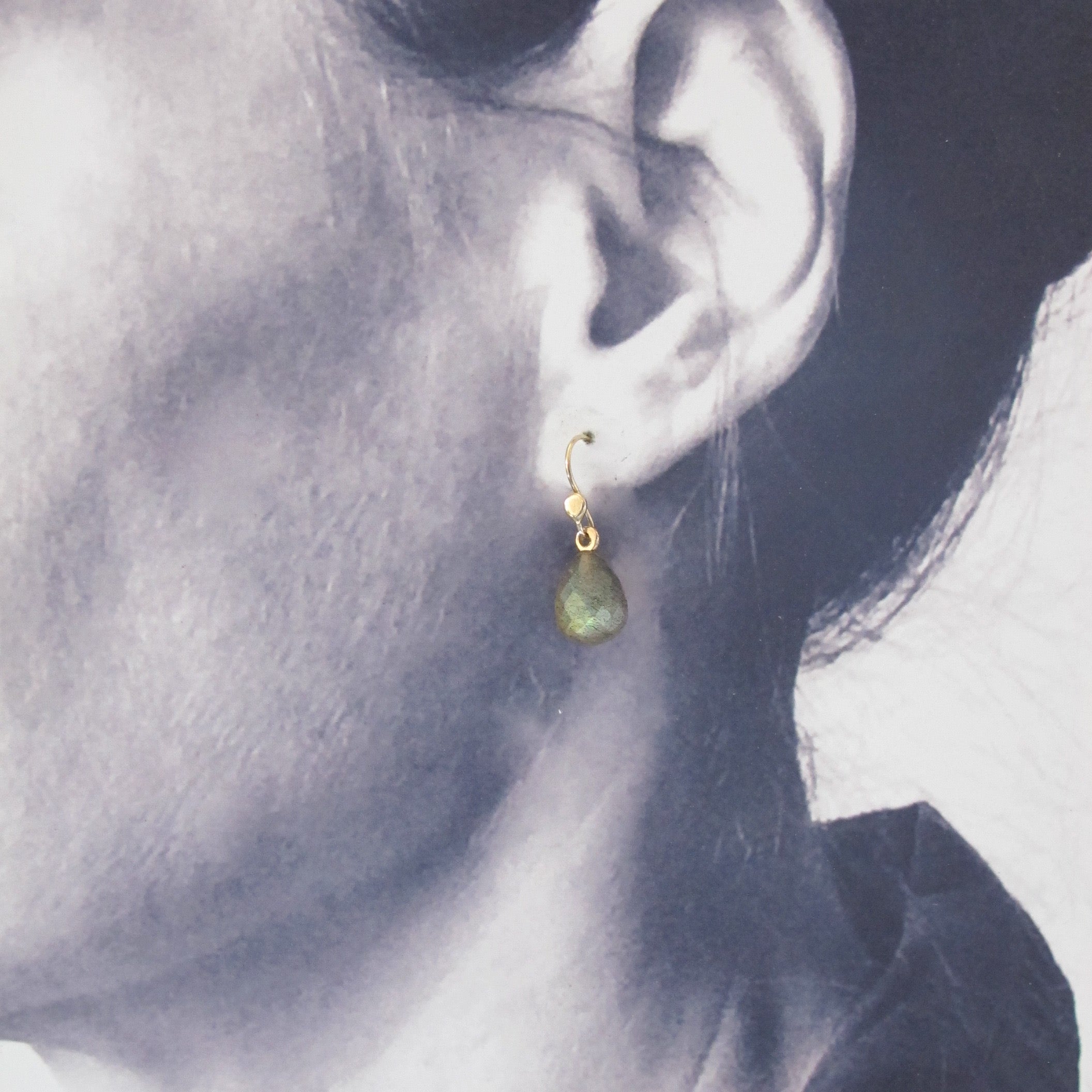 A close-up side profile of a person wearing a small gold hoop earring with a teardrop-shaped green stone dangle.