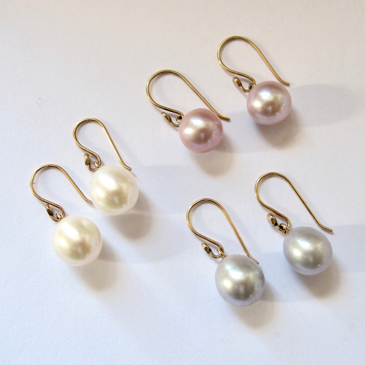 Five pearl drop earrings with gold French hook findings, shown in white, pale pink, and pale silver-gray on a plain white background.