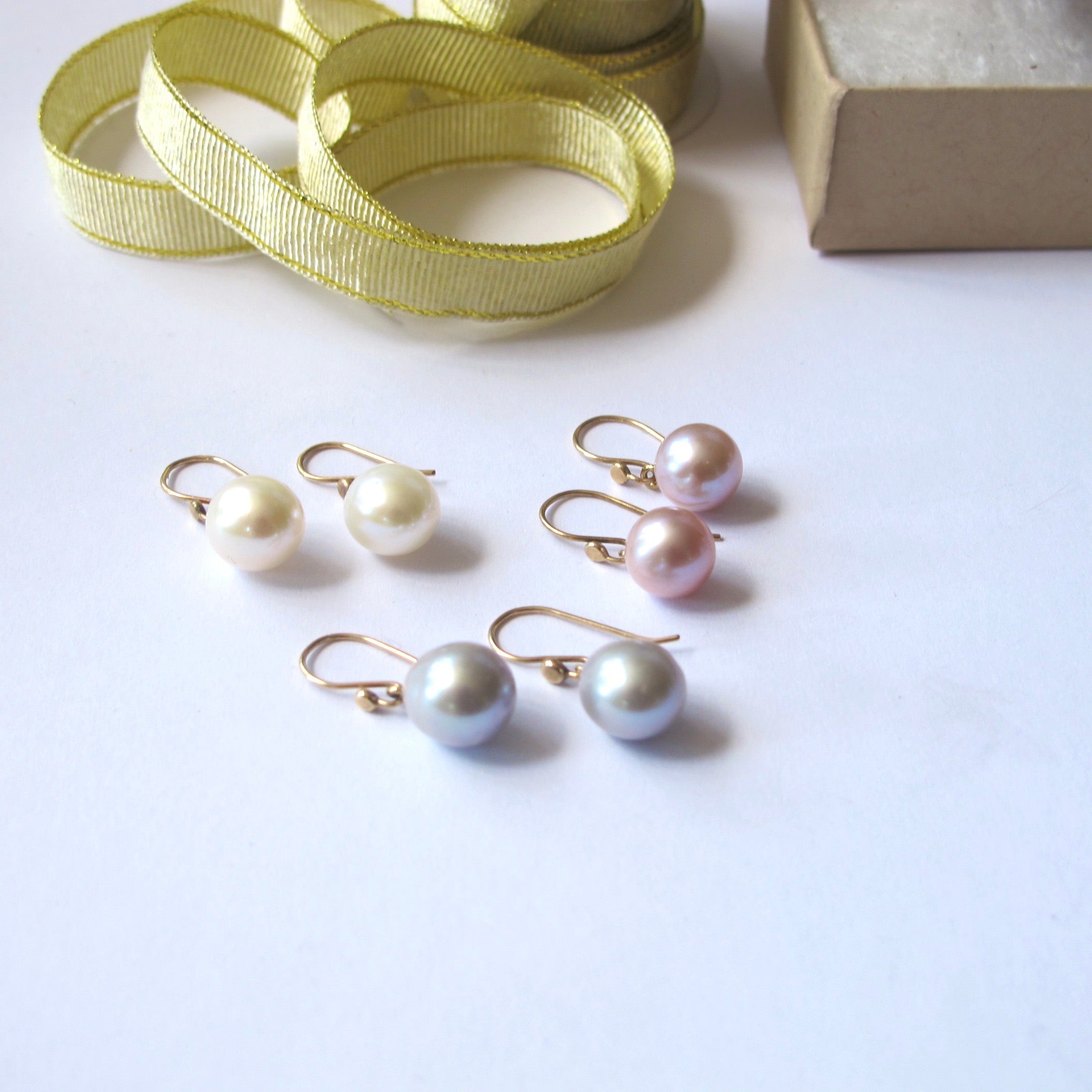 Four pairs of freshwater pearl drop earrings in white, pink, and silver-gray, each with gold fishhook wires, arranged on a white surface with a gold ribbon and a brown box in the background.