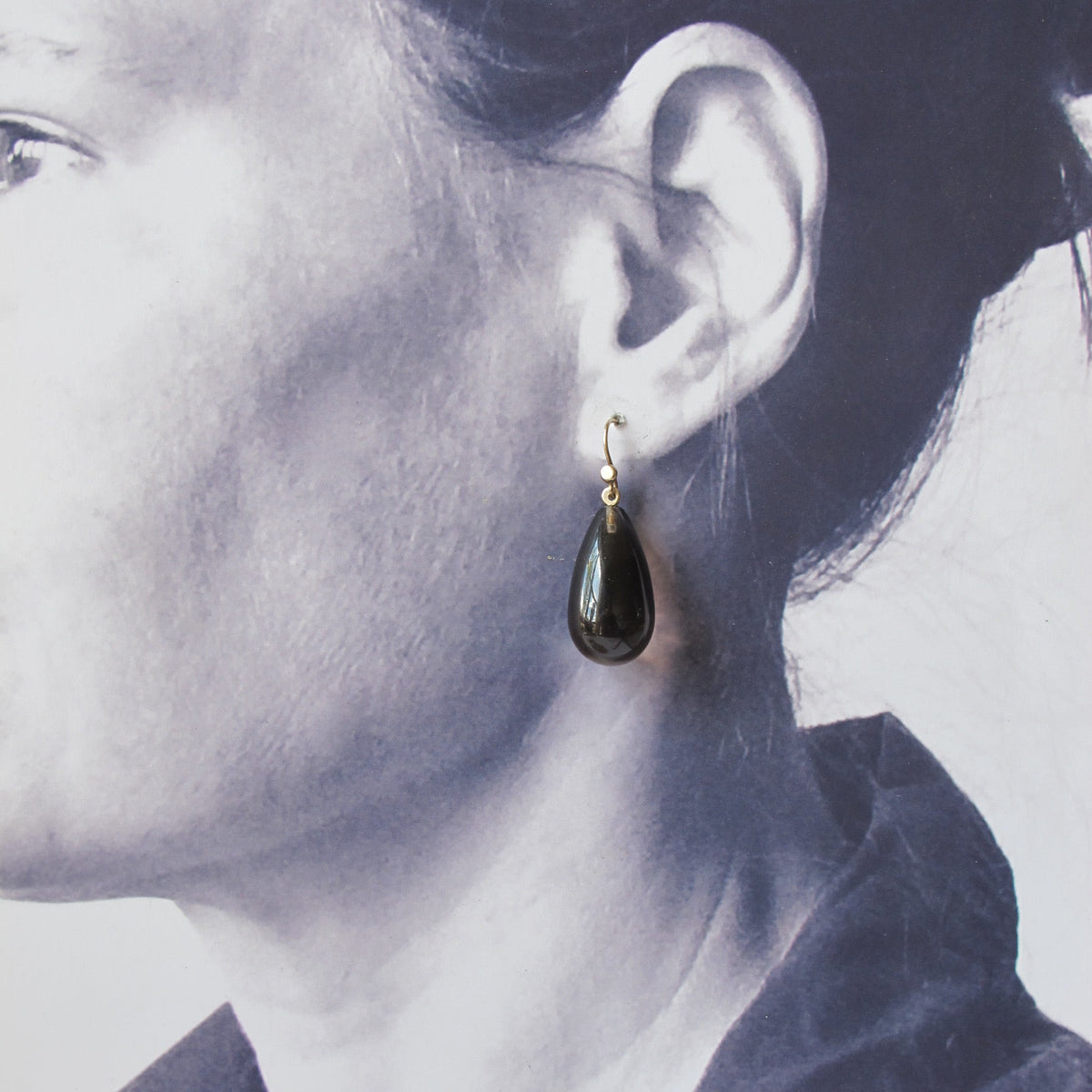 A close-up side profile of a woman wearing a single dark teardrop-shaped drop earring with a small gold hook.