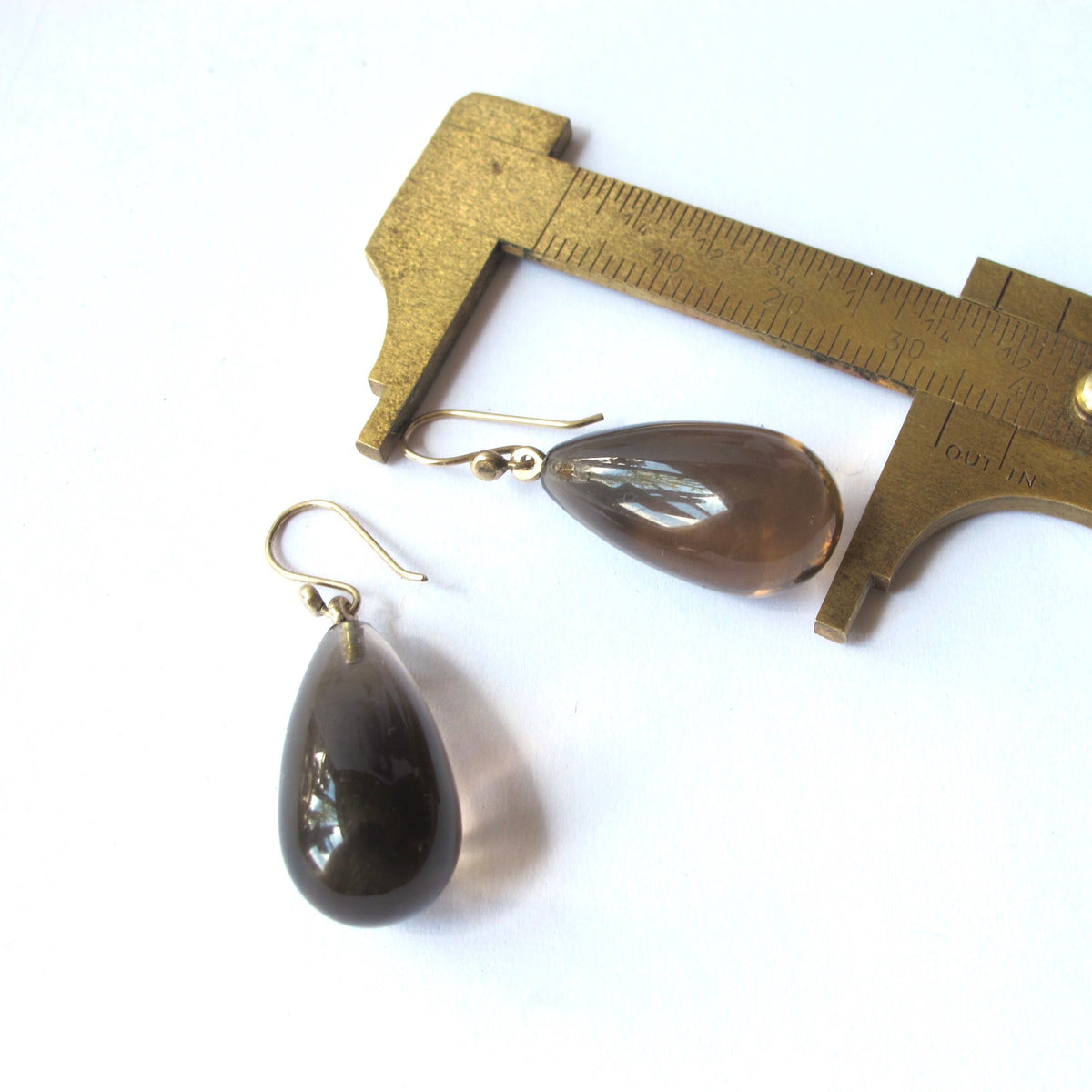 A pair of teardrop-shaped dark brown glass drop earrings with gold fishhook wires, displayed next to a brass caliper on a white background.
