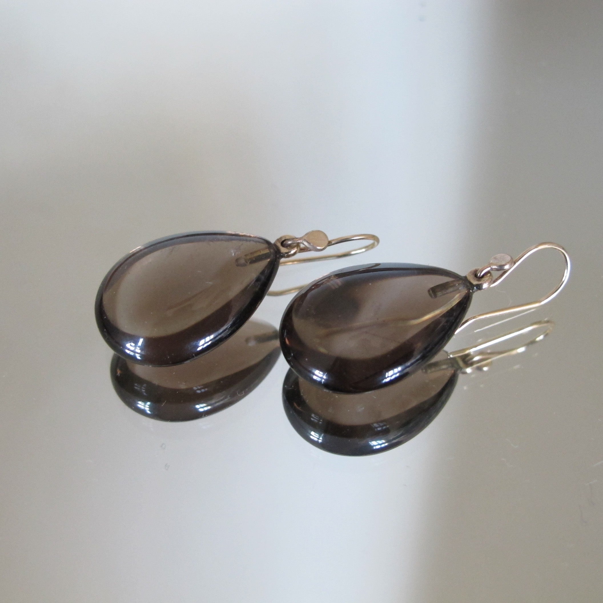 A pair of teardrop-shaped smoky brown glass drop earrings with gold-tone French hook ear wires, displayed on a reflective light beige surface.