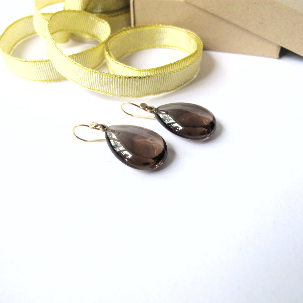 Teardrop-shaped smoky brown glass drop earrings with gold fishhook wires, displayed on a white surface next to a roll of yellow ribbon and a brown kraft paper box.