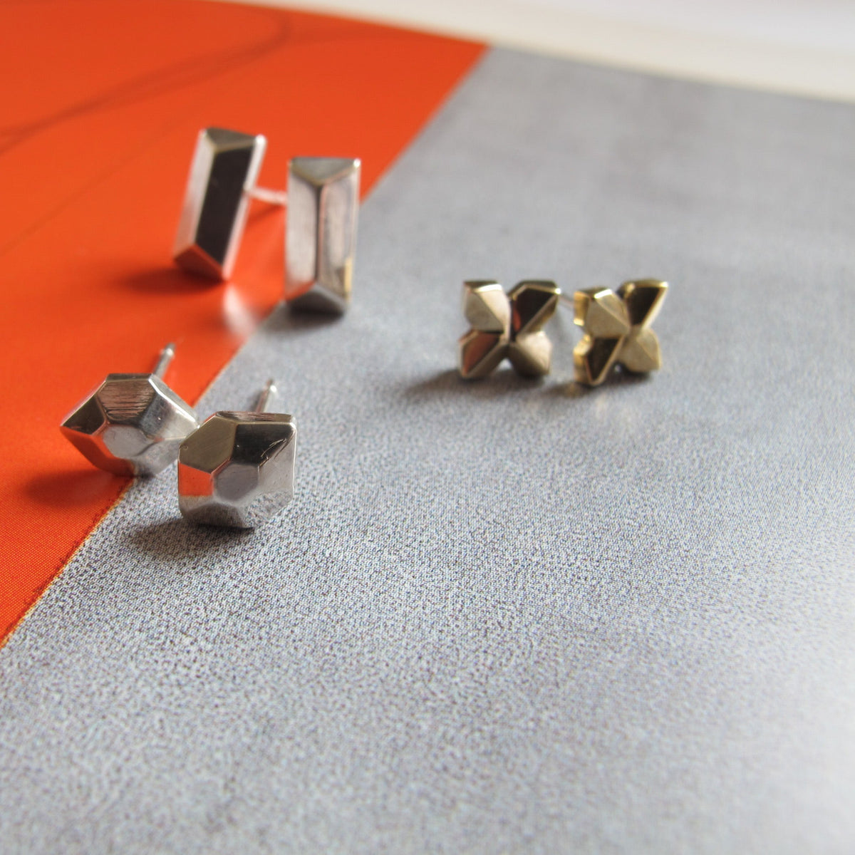 Four geometric stud earrings arranged on a split orange and gray surface. Two earrings are silver-toned and two are gold-toned, with one pair standing upright and the other lying flat.