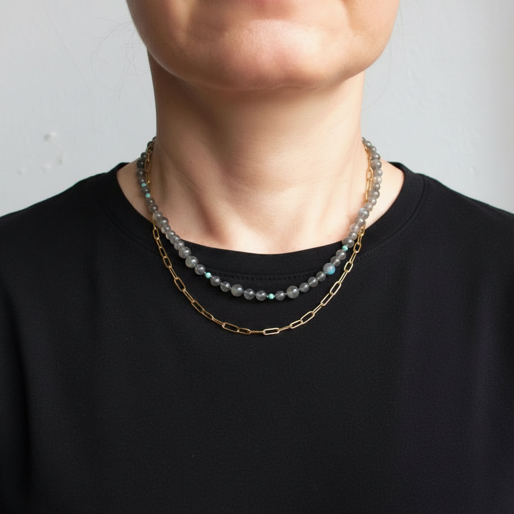 A close-up of a person's neck and lower face wearing two layered necklaces: one gold chain and one beaded necklace with gray, blue, and iridescent beads.