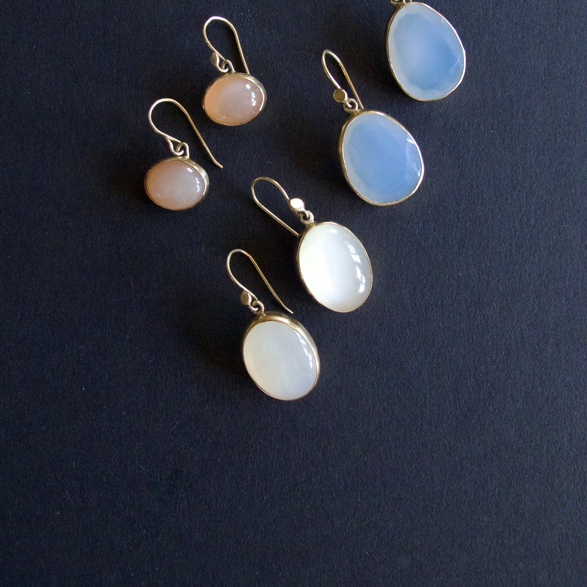 Five gold-tone drop earrings with oval and round cabochon gemstones in pale blue, white, and peachy-pink tones, arranged on a dark textured background.