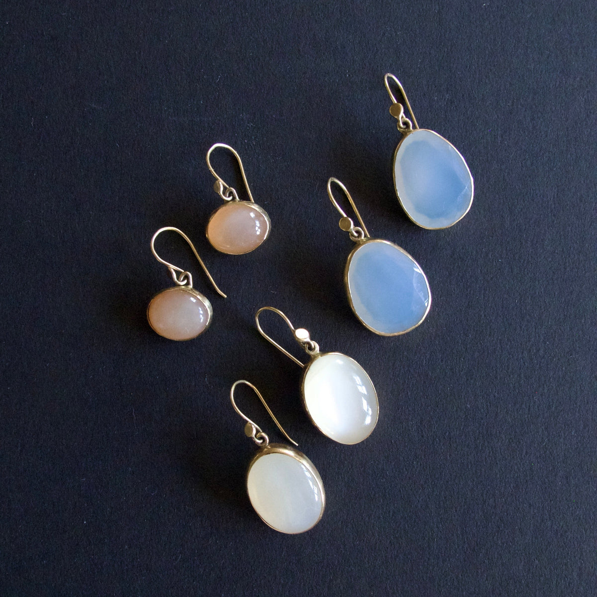 Five gold-tone drop earrings with oval and round cabochon gemstones in pale blue, white, and peachy-pink tones, arranged on a dark textured background.