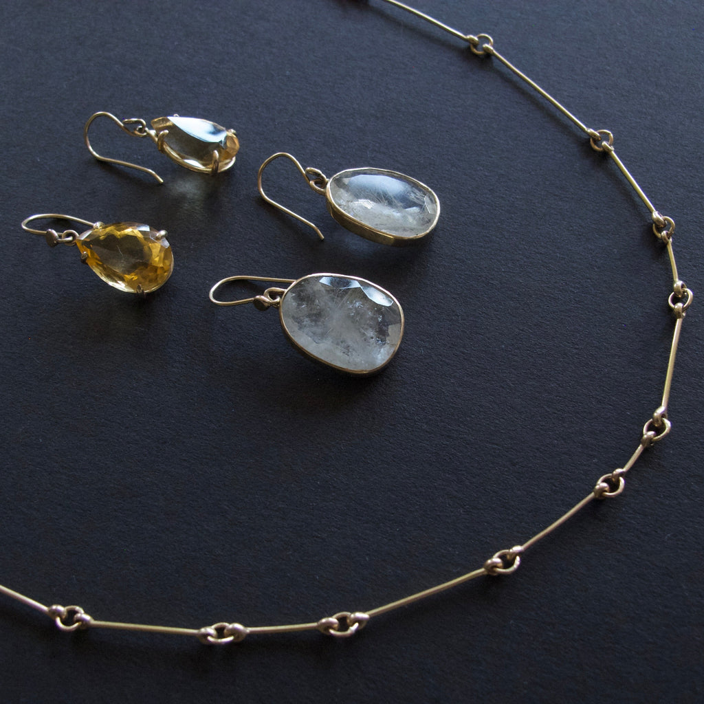 A jewelry set featuring a gold chain necklace and matching drop earrings with raw, polished gemstone pendants, displayed on a dark textured background.