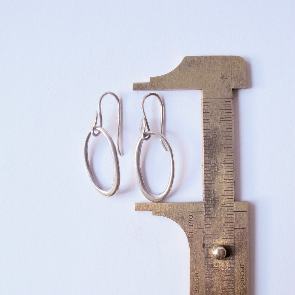 A pair of silver-toned oval drop earrings displayed next to a brass caliper on a plain white background, showing scale.