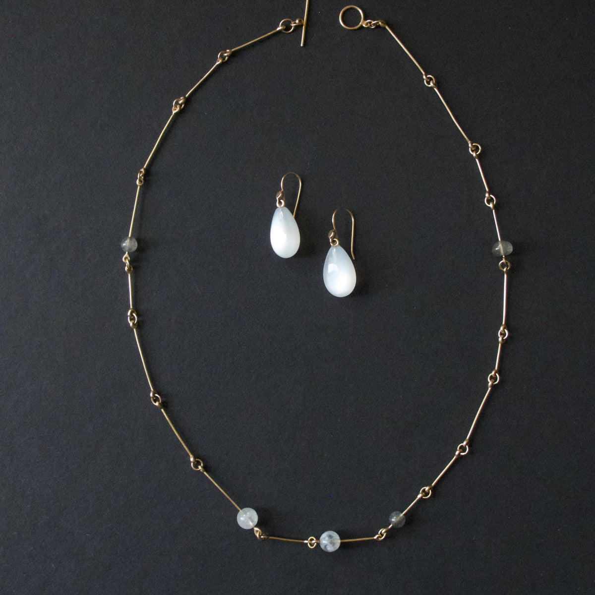 A gold-toned beaded necklace and matching drop earrings with white teardrop beads, arranged on a dark charcoal background.