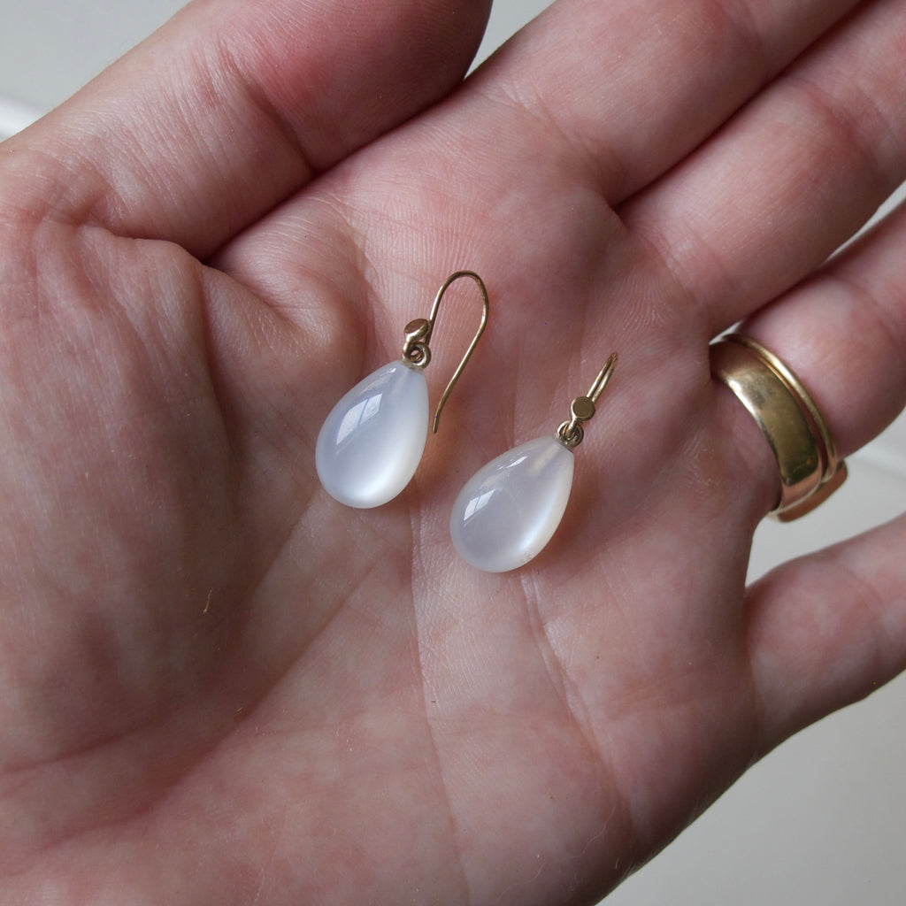 A pair of teardrop-shaped white pearl drop earrings with gold fishhook wires, held in the palm of a hand.