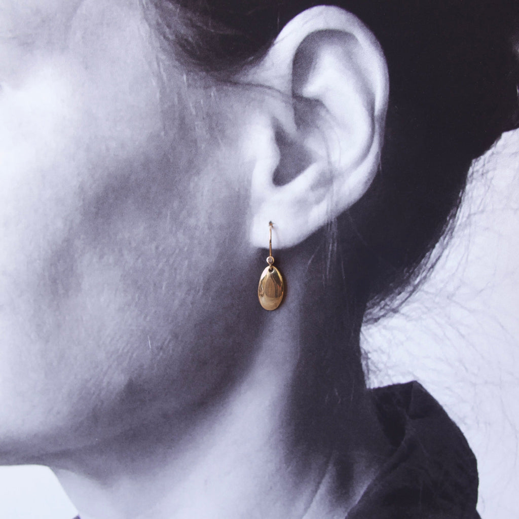 A close-up side profile of a woman's ear wearing a small gold teardrop dangle earring, shown in a high-contrast black and white photograph with a cool blue tint.