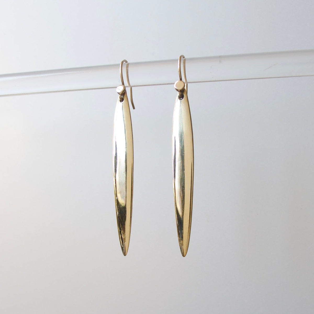 A pair of gold-toned, elongated teardrop-shaped dangle earrings hanging from a clear horizontal bar against a light gray background.