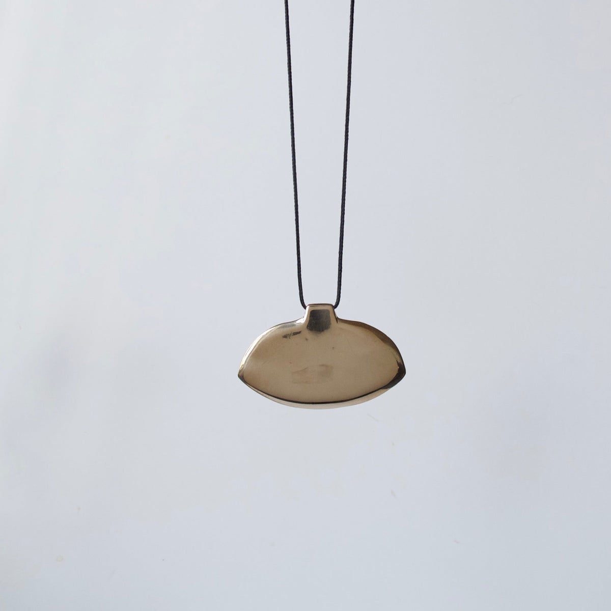 A minimalist pendant necklace featuring a small, organic-shaped metallic gold pendant suspended from a thin black cord, set against a plain light gray background.