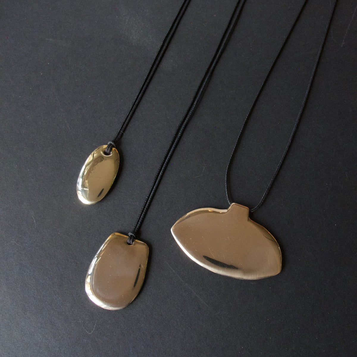 Two minimalist pendant necklaces with organic, fluid shapes in a brushed gold finish, hanging from thin black cords against a dark gray background.