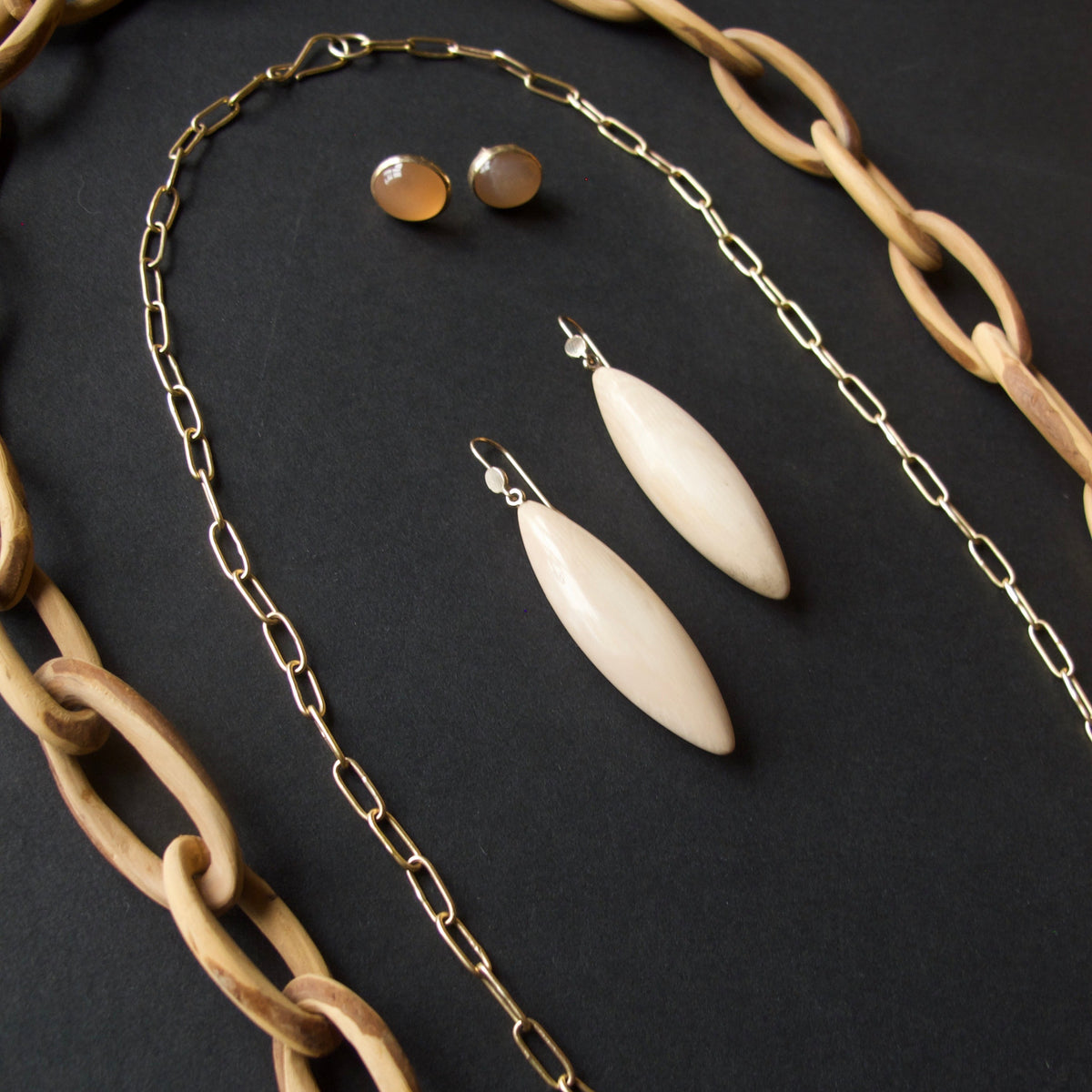 A jewelry set featuring a gold paperclip chain necklace, a gold link chain necklace, matching gold stud earrings, and two cream-colored teardrop-shaped drop earrings, arranged on a dark textured background with a wooden chain partially framing the shot.