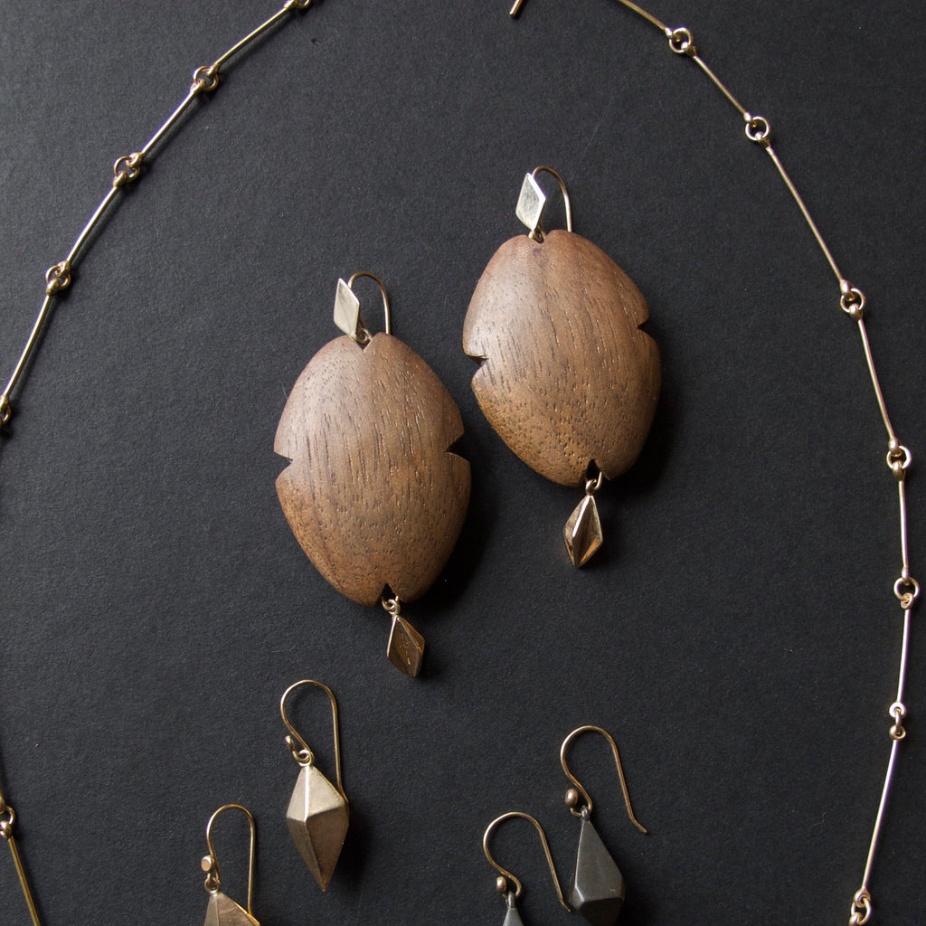 A jewelry set displayed on a dark background, featuring a delicate gold beaded necklace, a pair of large oval wooden drop earrings, and several smaller geometric gold and dark gray drop earrings.