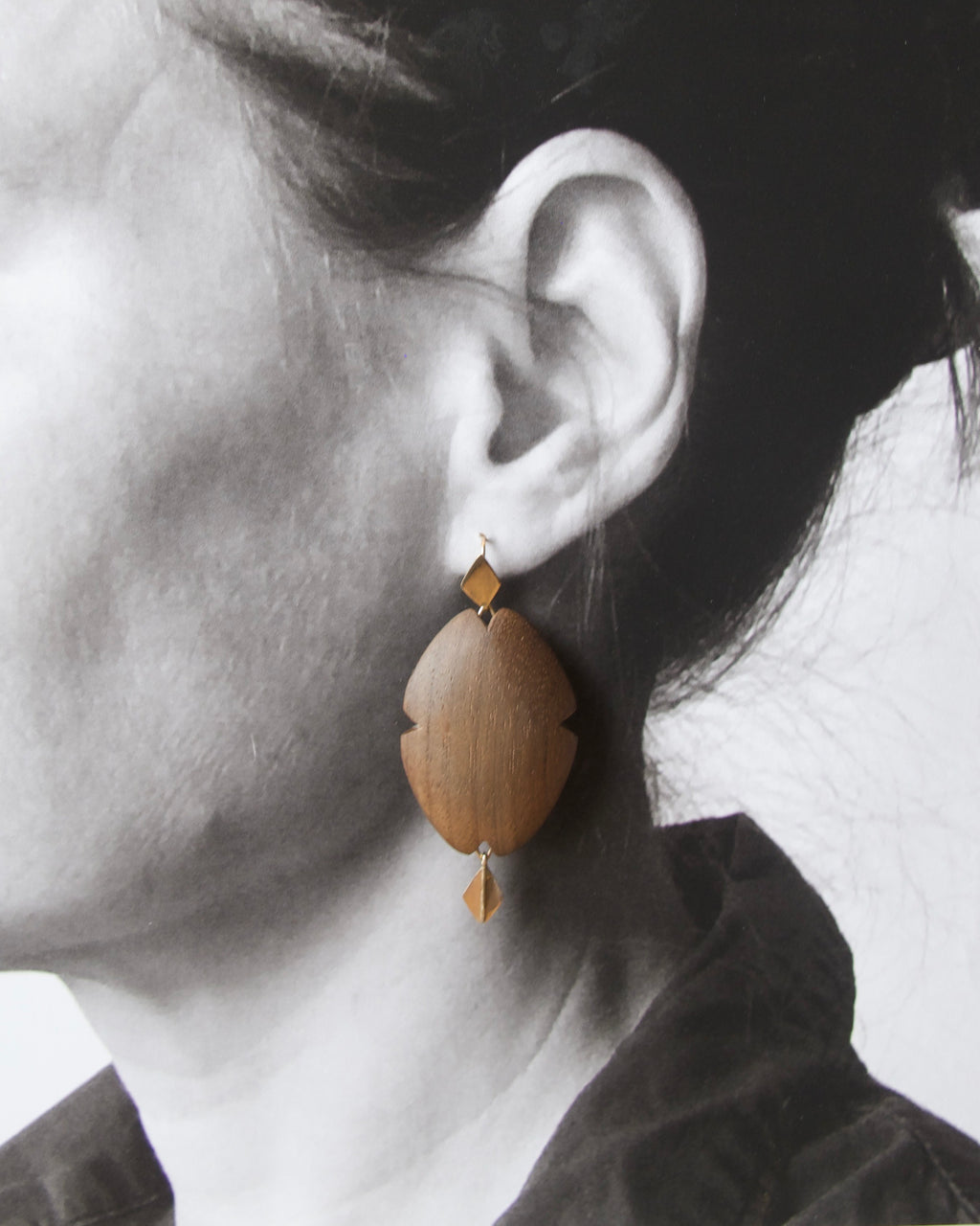 A close-up of a person's ear wearing a single organic-shaped wooden dangle earring with small geometric gold-toned accents.
