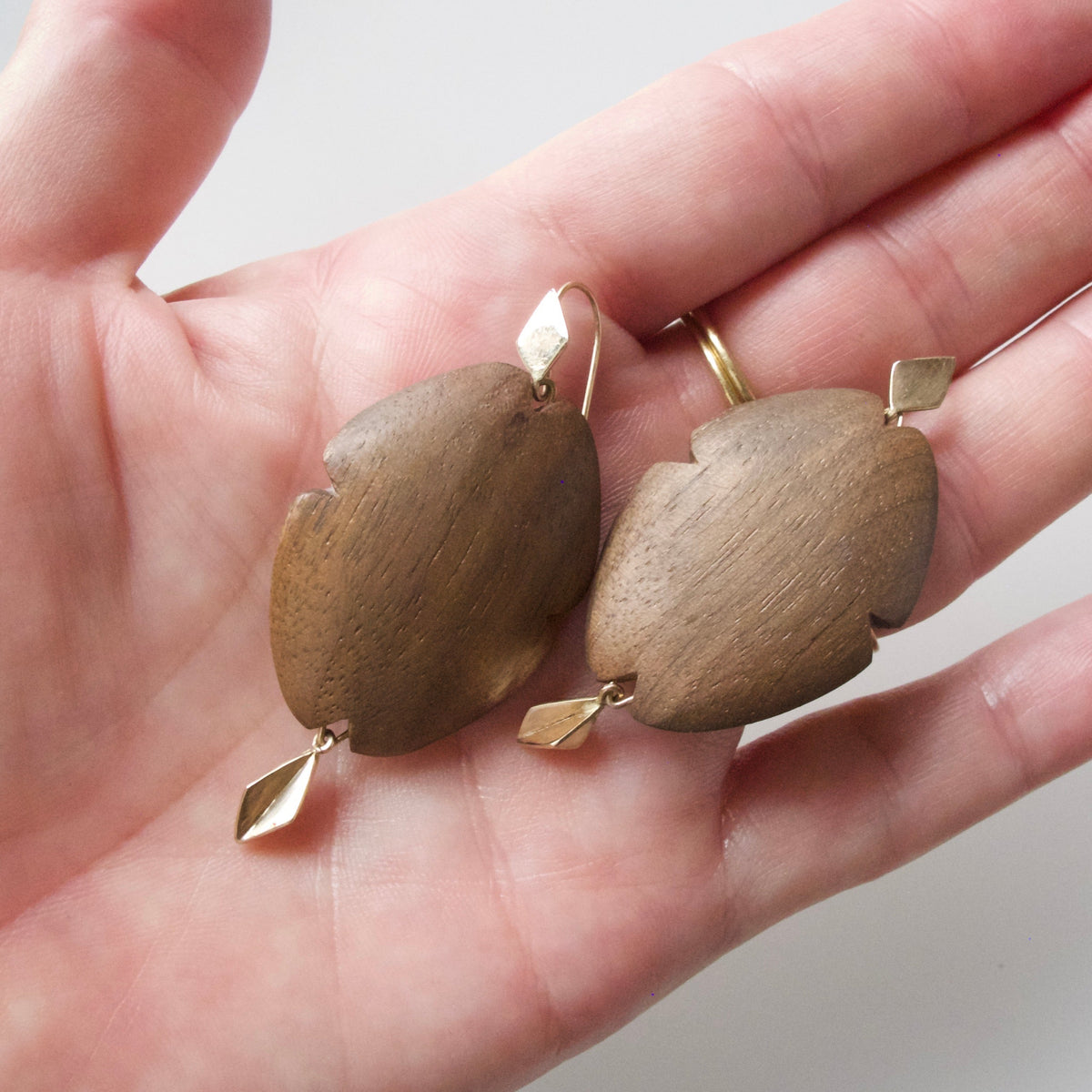 A pair of organic-shaped wooden drop earrings with gold-toned metal hooks and small dangling gold accents, held in a person's hand against a light neutral background.