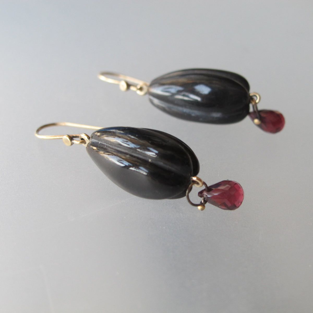 A pair of drop earrings featuring polished black teardrop-shaped beads suspended from gold hooks, with small dangling red teardrop gemstones, shown on a neutral light gray background.