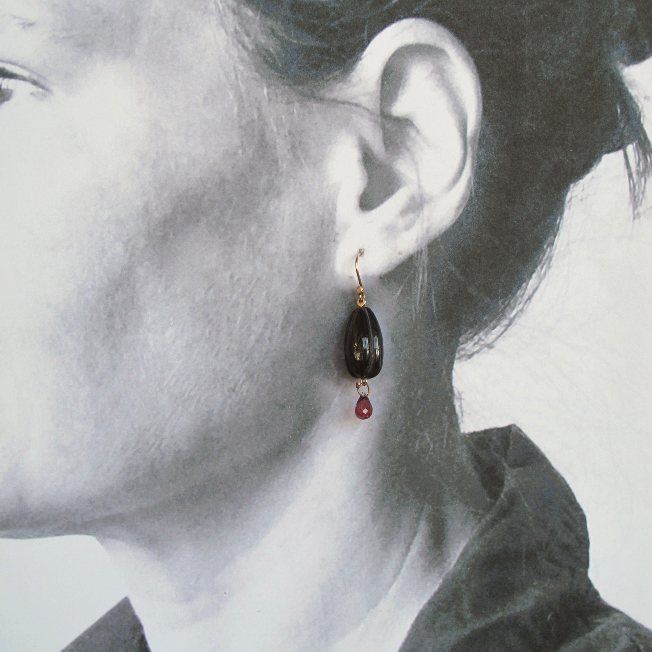 A close-up side profile of a woman wearing a dangling earring. The earring has a gold-toned fishhook and top cap, a dark oval stone, and a small red teardrop charm below.