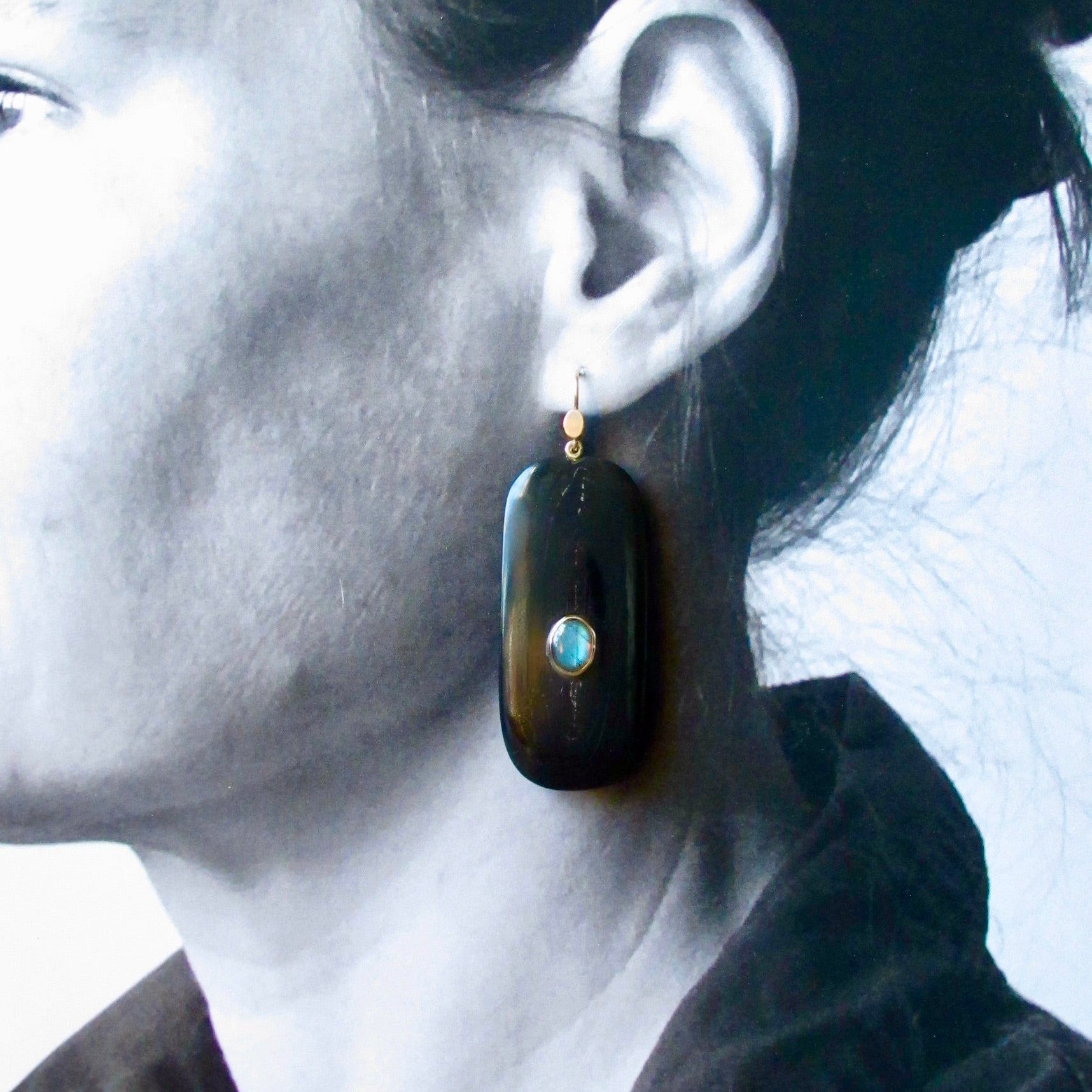 A close-up side profile of a woman wearing a dark elongated drop earring with a small turquoise-blue stone and gold-tone setting.
