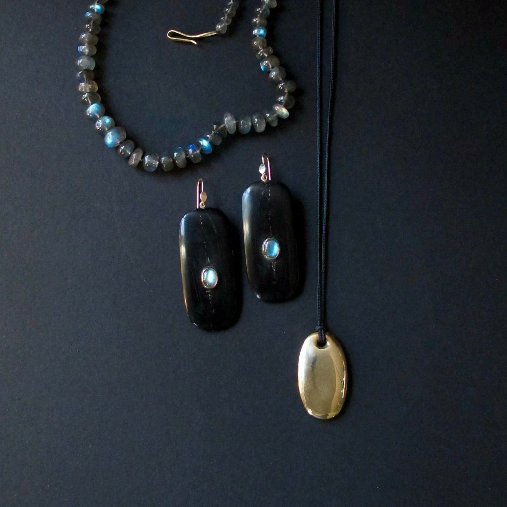 A jewelry set featuring a beaded necklace with gray and blue-green beads, a pair of black rectangular drop earrings with light blue stone accents, and a gold oval pendant on a black cord, arranged on a dark background.