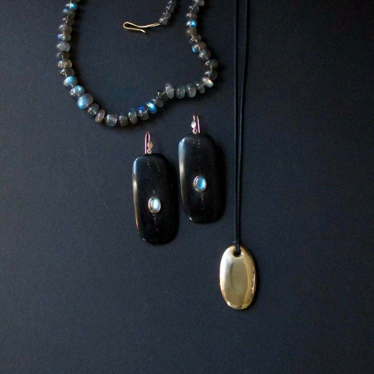 A jewelry set featuring a beaded necklace with gray and blue-green beads, a pair of black rectangular drop earrings with light blue stone accents, and a gold oval pendant on a black cord, arranged on a dark background.