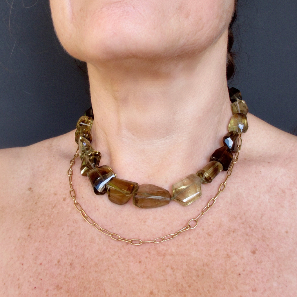 A close-up of a person's neck and upper chest wearing a chunky gold-toned beaded necklace with faceted brown and smoky amber stones, layered over a delicate gold chain.