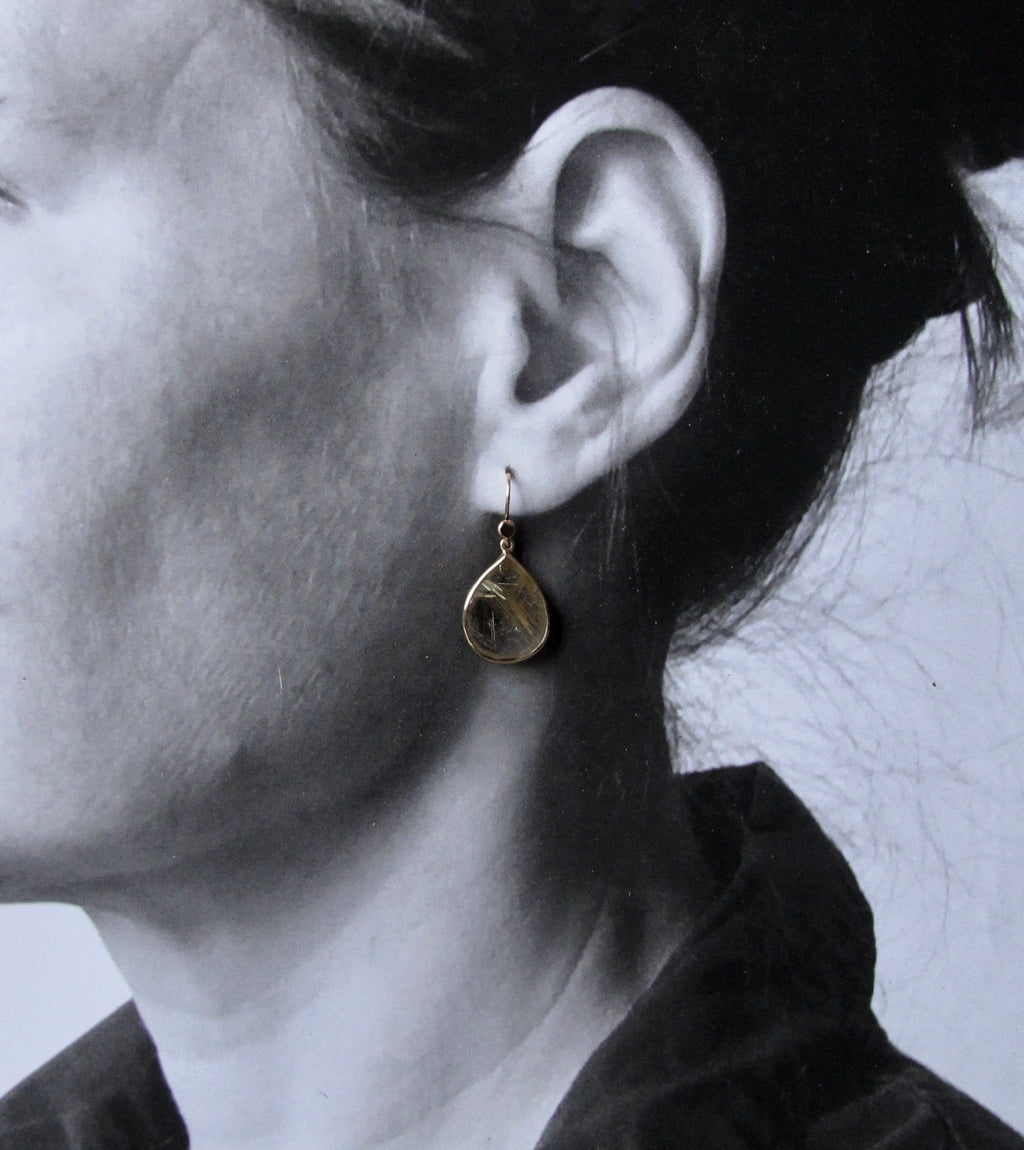 A close-up side profile of a person wearing a small teardrop-shaped gold pendant earring with a textured, hammered surface.