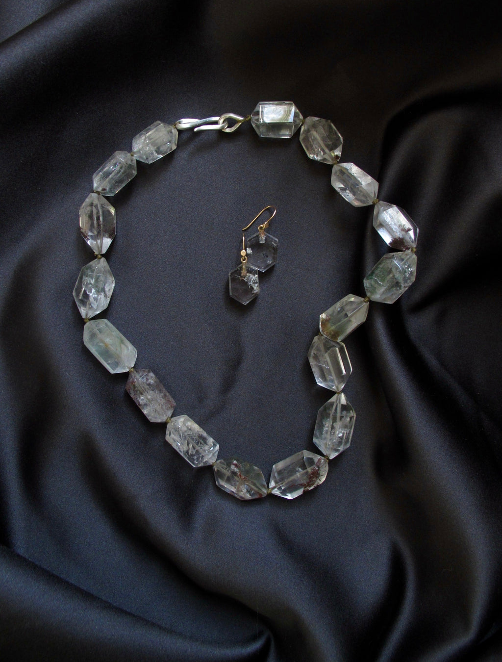 Faceted clear gemstone beaded necklace and matching drop earrings displayed on a black satin fabric background.