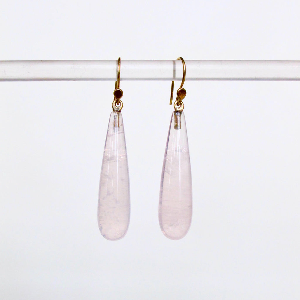 A pair of teardrop-shaped light pink gemstone dangle earrings with gold hooks, hanging from a clear horizontal display bar against a plain white background.