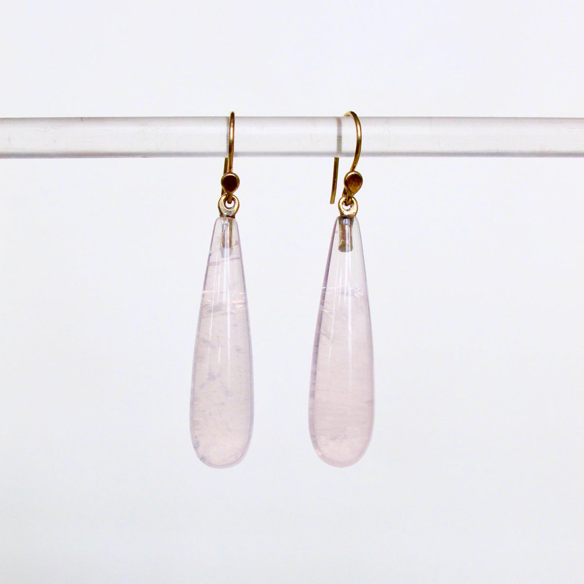 A pair of teardrop-shaped light pink gemstone dangle earrings with gold hooks, hanging from a clear horizontal display bar against a plain white background.
