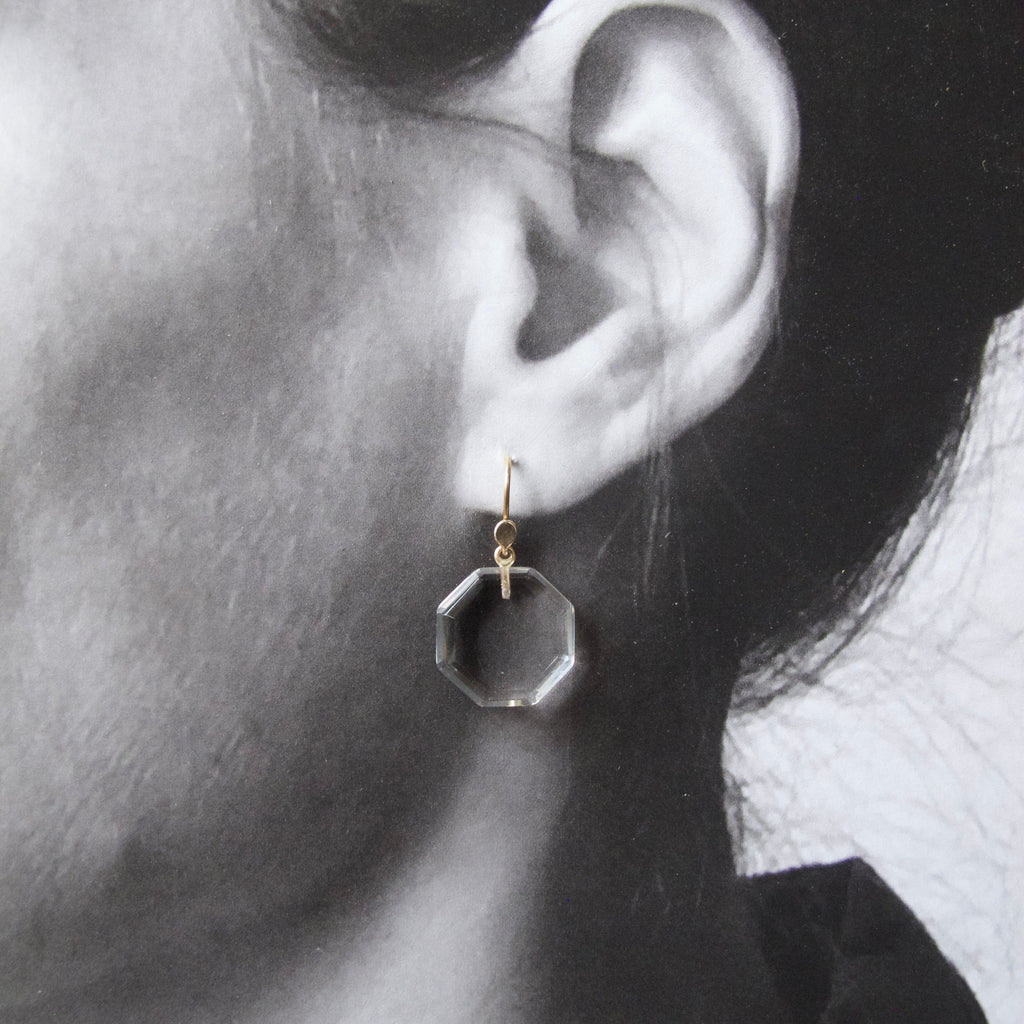 A close-up of a person's ear wearing a small geometric dangling earring with a gold hook and a clear faceted octagonal drop.