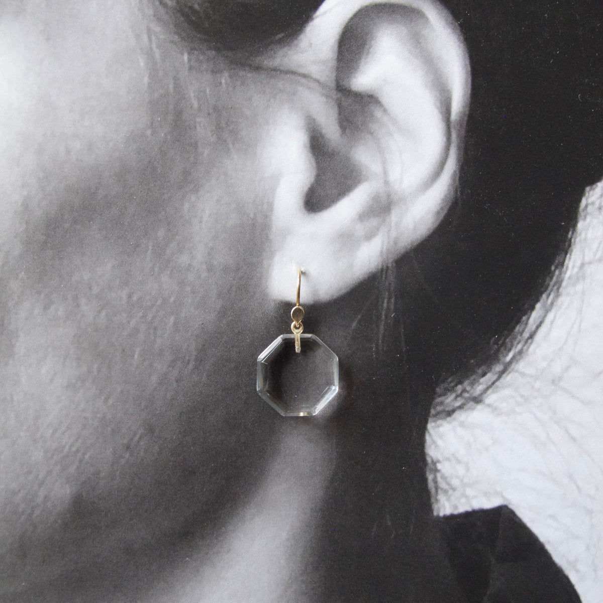 A close-up of a person's ear wearing a small geometric dangling earring with a gold hook and a clear faceted octagonal drop.