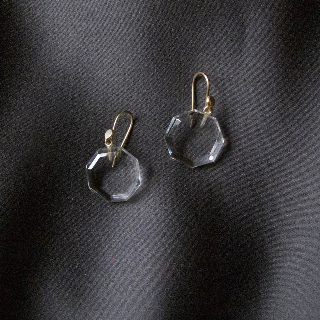 A pair of clear faceted octagonal drop earrings with gold fishhook wires, displayed on a dark, textured satin fabric background.